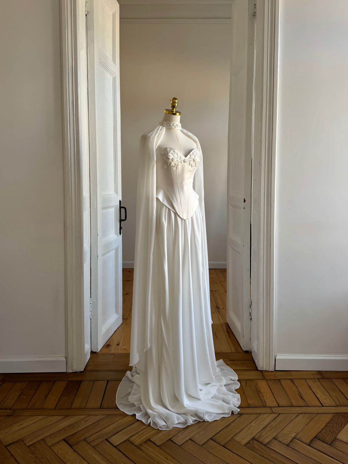 White wedding dress on a mannequin with a long train in a room with wooden flooring and white walls.